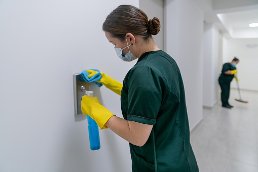 Latin American maid wearing a facemask while cleaning the buttons of an elevator at an office building during the COVID-19 pandemic