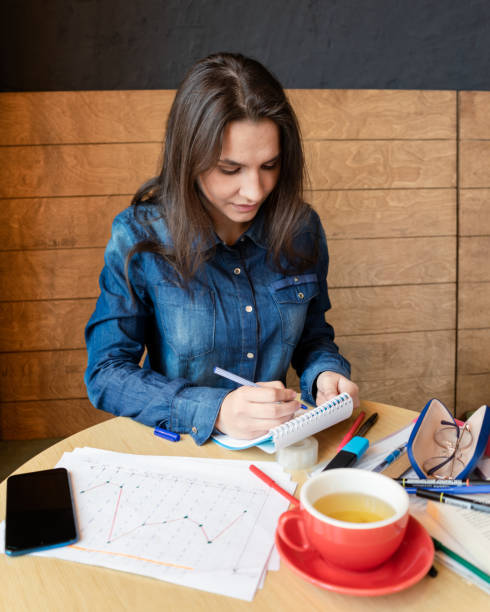 Girl in a blue denim shirt sitting in a cafe makes notes in a notepad with a pen. A smartphone, sheets with various charts, glasses in a case, a red mug with green tea, felt-tip pens are on the table una chica con una camisa de mezclilla azul sentada en un café hace notas en un bloc de notas con un bolígrafo. un teléfono inteligente, sábanas con varios gráficos, gafas en una caja, una taza roja con té verde, rotuladores están en la mesa