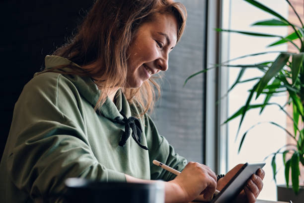 istock Long-haired woman draws on a tablet at cafe table in front of large window. Selective focus 1312491094