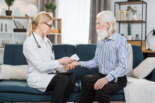 istock Middle aged practitioner visiting sick elderly man patient at home. Young blond woman doctor measuring patient temperature with non-contact wrist infrared thermometer at home. 1312502562