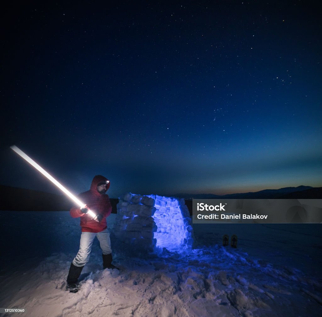 Jedi concept. Tourist using light near igloo at night while camping in winter mountain. Alone in nature while COVID-19 pandemic. Mental health and life balance. Man spending vacation in the nature during coronavirus pandemic. Using technologies  to talk online with friends. Laser Sword Stock Photo