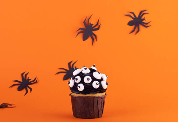 black cupcake with lots of marshmallow eyes for halloween on an orange background with black spiders black cupcake with lots of marshmallow eyes for halloween on an orange background with black spiders. mystical scary background halloween cupcake stock pictures, royalty-free photos & images