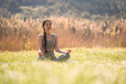 Young woman sitting in the grass while meditating in the wilderness. Vertical