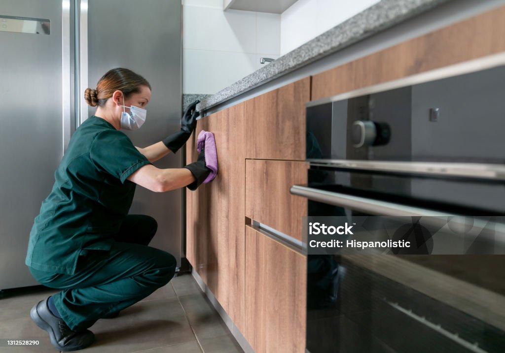 Latin American cleaner wearing a facemask while cleaning the kitchen Latin American cleaner wearing a facemask while cleaning the kitchen at a house during the COVID-19 pandemic Housework Stock Photo