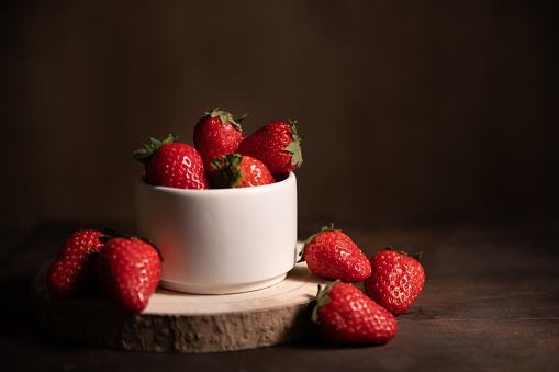 Ripe garden strawberry in white ceramic bowl on dark table. Fresh strawberries on brown wooden background. Healthy food. Selective focus, strong shadows, dark photography style.