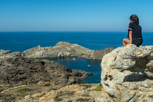 Rear view of a teenage girl sitting on the rocks of the Cap de Creus (Cape of Crosses) headland in Catalonia, Spain. The young traveler observes the sea and a small boat in the bay. Moments of relaxation, inner reflection and concept of freedom.