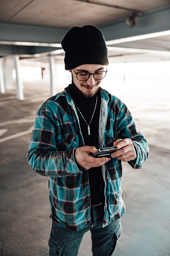 Happy smiling young man checking social media news and messages on his mobile phone standing inside urban parking garage. Millennial Generation Real People Social Media Lifestyle.