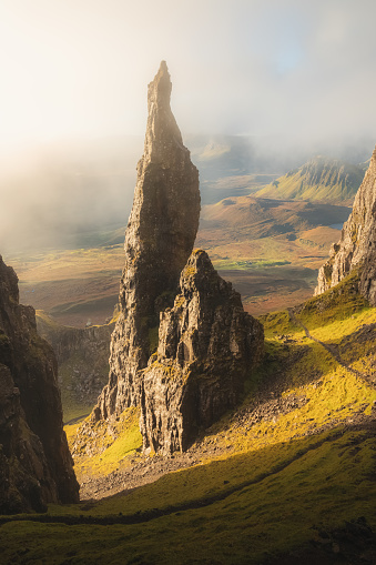 Moody, atmospheric fog and mist with golden sunrise or sunset light and shadow at the iconic Needle rock pinnacle at the Quiraing on the Trotternish Ridge, Isle of Skye.