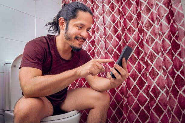 istock Young latin man smiling on the toilet using a smart phone at the bathroom 1312557848
