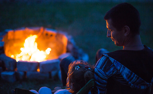 Dad and daughter sit at night by the fire in the open air in the summer in nature. Family camping trip