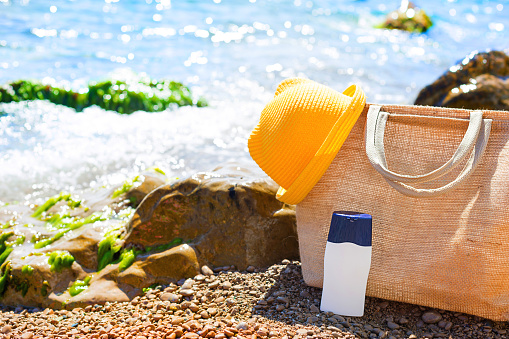 Beach bag with a white tube of sunscreen on a pebble beach near the sea. Travel, beach holiday at the resort, glasses, yellow hat. Protection of the skin from UV rays, suntan oil. Copy space, mock up.