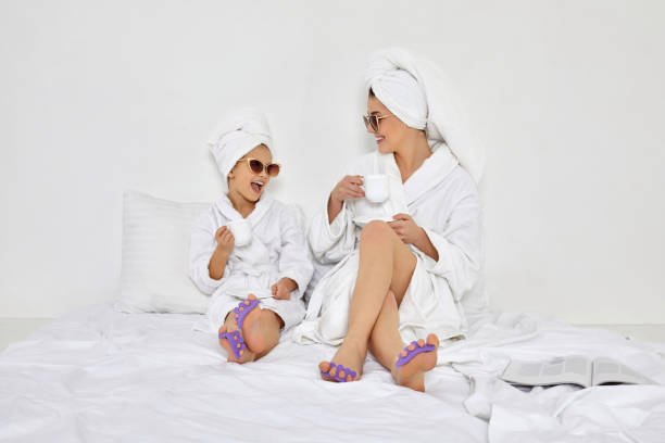 happy woman and her daughter in white bathrobes glamorous woman and her daughter in white bathrobes and towels on heads with pedicure sponge on fingers drinking tea. mom spending time together with kid towel photos stock pictures, royalty-free photos & images