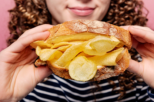 Close-up cropped portrait of a young woman holding a staple of the english diet a “Crisp Sandwich”. Potato chips between two slices of buttered white bread, a snack, a meal a hangover cure, you decide! Delicious. Colour, horizontal with some copy space.