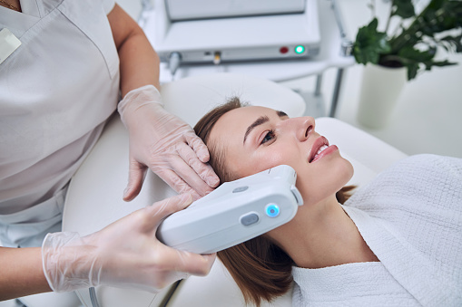 Close up side view portrait of young cheerful female lying on medical chair while unrecognized doctor making non surgical face lifting in beauty salon
