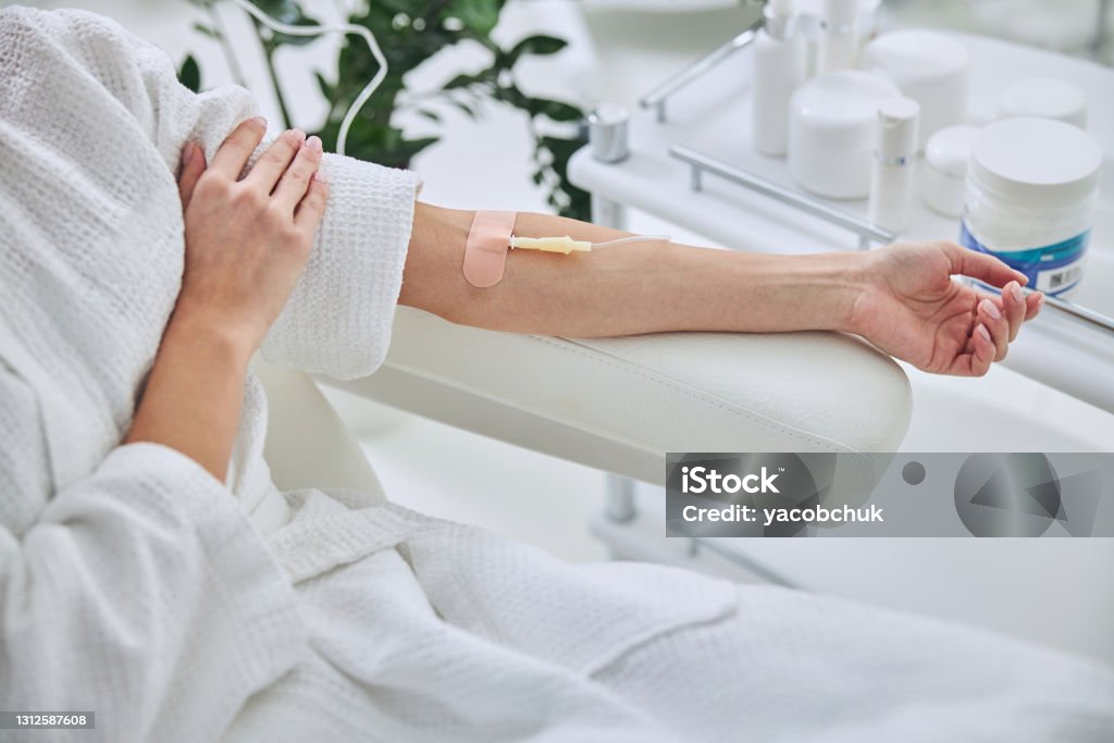 Young female in white bathrobe during medical procedure in beauty clinic Cropped head portrait of unrecognized female patient sitting in medicine armchair while receiving IV infusion in wellness center IV Drip Stock Photo