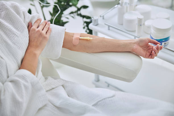 Young female in white bathrobe during medical procedure in beauty clinic Cropped head portrait of unrecognized female patient sitting in medicine armchair while receiving IV infusion in wellness center alternative therapy stock pictures, royalty-free photos & images