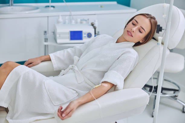 Redheaded beautiful female resting and getting IV infusion in spa salon Waist up side view portrait of young pretty lady in white bathrobe lying on medicine armchair while receiving vitamin medical procedure in wellness center infused stock pictures, royalty-free photos & images