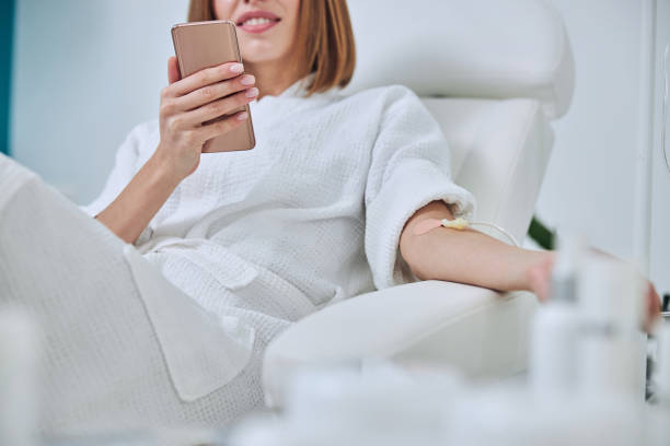 Beautiful Caucasian woman sitting in armchair during medicine procedure in spa salon Cropped head portrait of unrecognized young lady in white bathrobe holding mobile phone in hand while receiving intravenous vitamin drip treatment in wellness center alternative therapy stock pictures, royalty-free photos & images