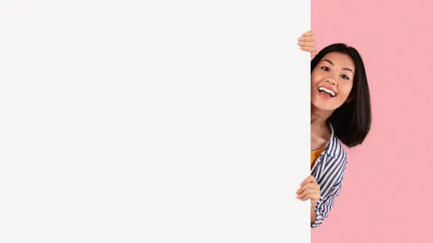 Intresting Offer. Happy Casual Asian Woman Peeping Out The Side Of White Advertisement Board For Your Text Or Design. Smiling Lady Holding Billboard, Looking At Camera, Standing Over Pink Wall