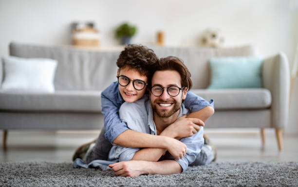 père et fils heureux ayant l’amusement à la maison. papa se trouvant sur le garçon de transport de tapis sur le dos et souriant ensemble à l’appareil-photo - glasses child little boys happiness photos et images de collection