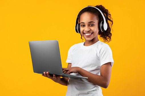 Kids And Technology Concept. Portrait of smiling black girl holding and using laptop computer, wearing wireless headphones, studying, watching online course, isolated over yellow studio wall
