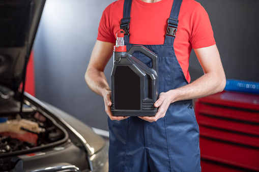 Car fluid. Hands of car mechanic in overalls holding black canister in workshop, face is not visible