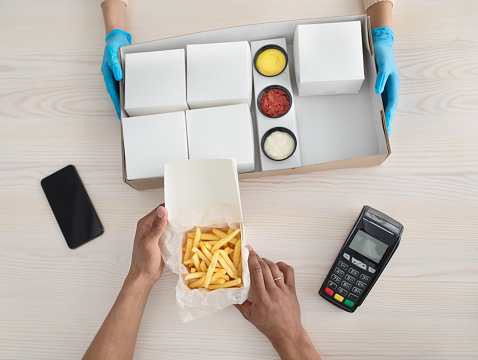 Covid-19 epidemic and safe fast food at home or office. Millennial african american man take box with fries, courier in rubber gloves holds order, near terminal, phone with blank screen, top view