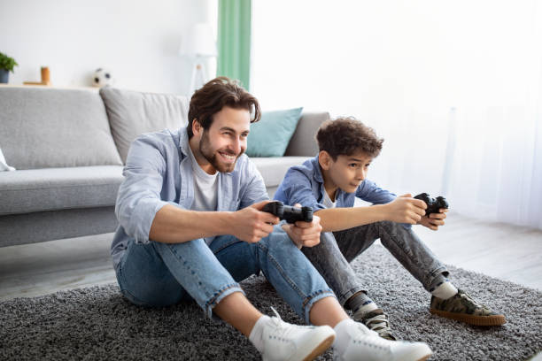 radosny ojciec i syn rywalizujący w internetowych grach wideo, używając joysticków, bawiąc się w domu. - video game friendship teenager togetherness zdjęcia i obrazy z banku zdjęć