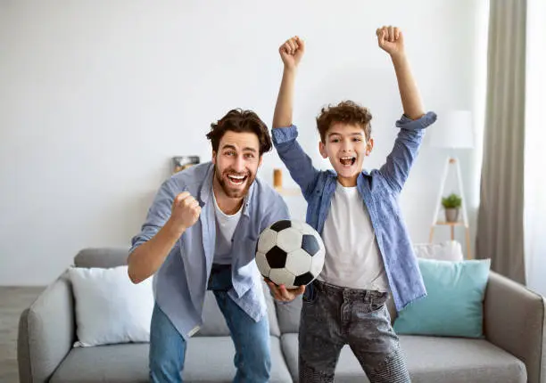 Two generations of football fans. Happy dad and son cheering soccer at home, emotionally supporting favorite team. Family watching sports on TV, celebrating goal with raised hands