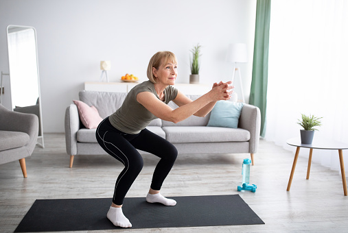 Positive senior lady doing squats on domestic workout in living room, free space. Fit mature woman leading active lifestyle, losing weight, staying in good shape at home during coronavirus quarantine