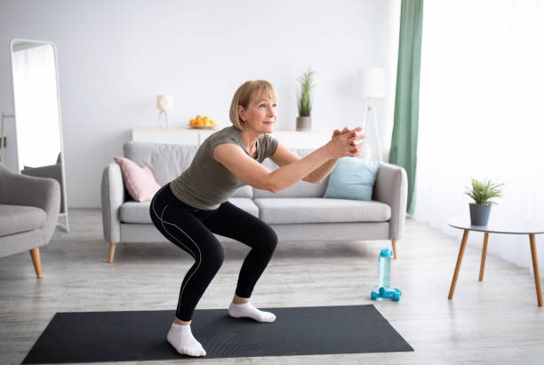 Positive senior lady doing squats on domestic workout in living room, free space Positive senior lady doing squats on domestic workout in living room, free space. Fit mature woman leading active lifestyle, losing weight, staying in good shape at home during coronavirus quarantine crouching stock pictures, royalty-free photos & images