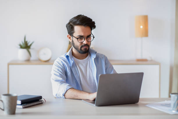 istock Telecommuting. Eastern Man Using Laptop Computer, Sitting At Desk In Home Office 1312608178