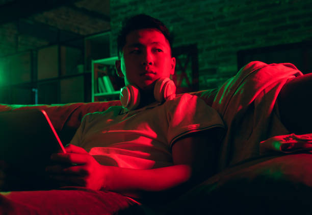 istock Young Korean man using tablet at home interior in gradient red green neon light 1312608404