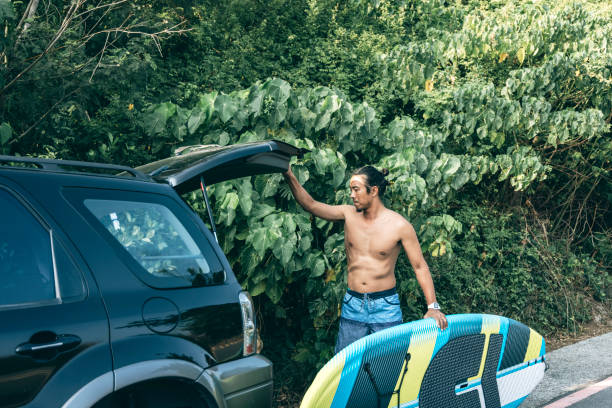 Back view of a man in a yellow hoodie holding a blue paddle board. Preparing for an outdoor adventure. Relaxing in a sunlit park with greenery and water views.