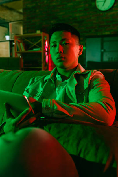 istock Young Korean man using tablet at home interior in gradient red green neon light 1312608553