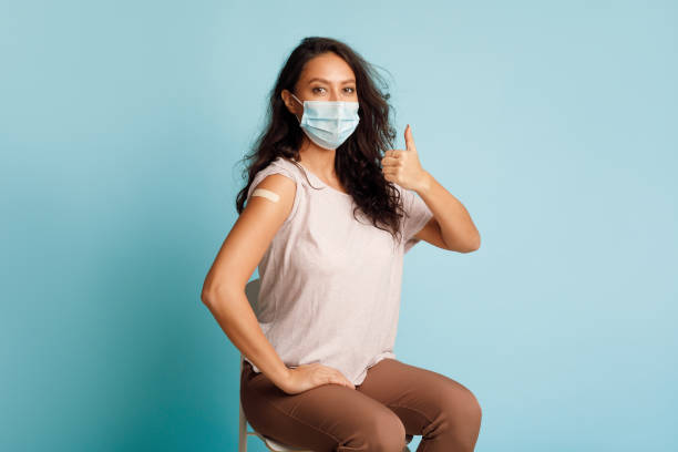 Vaccinated Woman Gesturing Thumbs-Up After Coronavirus Vaccination On Blue Background Covid-19 Vaccination. Vaccinated Woman Gesturing Thumbs Up Showing Arm With Plaster Bandage After Coronavirus Vaccine Injection On Blue Background, Wearing Protective Face Mask. Antivital Immunization adhesive bandage photos stock pictures, royalty-free photos & images