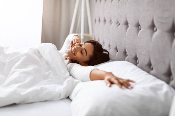 Satisfied young black woman stretching after wake up Good Start Of Sunny Day. Portrait of well-slept positive black lady stretching arms after waking up, feeling lazy, happy beautiful woman lying with closed eyes in bed covered with white blanket person waking up stock pictures, royalty-free photos & images