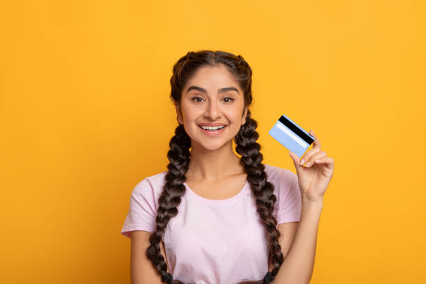 mujer india sonriente sosteniendo tarjeta de crédito, pared amarilla del estudio - greeting card holding women credit card fotografías e imágenes de stock