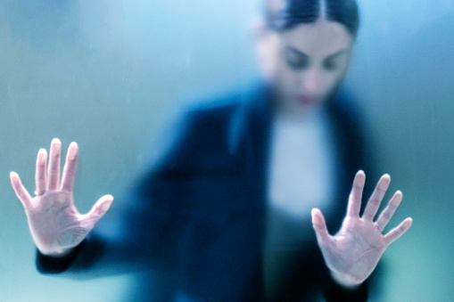 Diffused silhouette of female gesturing fear behind frosted glass