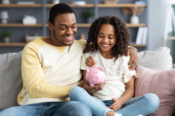 Black girl and father putting coin into piggy bank Black girl and father putting coin into piggy bank, sitting on sofa at home. Happy african american dad teaching his cute daughter how to save money, living room interior. Financial education concept financial literacy stock pictures, royalty-free photos & images