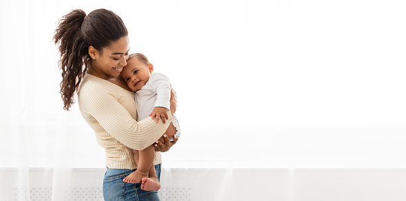 Smiling African Mother Holding Baby Boy In Arms Hugging And Cuddling Cute Little Infant Standing Near Window Indoors. Child Care, Happy Motherhood Concept. Panorama With Empty Space For Your Text