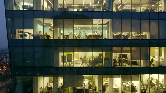 Aerial view of the skyscraper's nighttime windows with office workers sitting at computers and conducting confirmations.