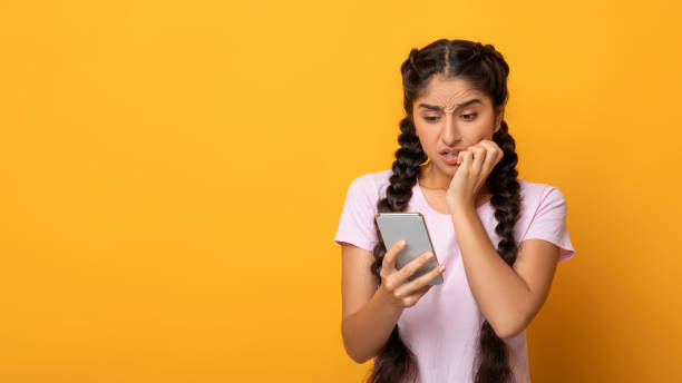 Worried scared indian woman looking at phone seeing bad news Bad News. Worried nervous indian young lady holding and looking at her cellphone,r eading negative comment. Afraid woman biting nails isolated on yellow studio background, banner, free copy space sad disbelief stock pictures, royalty-free photos & images