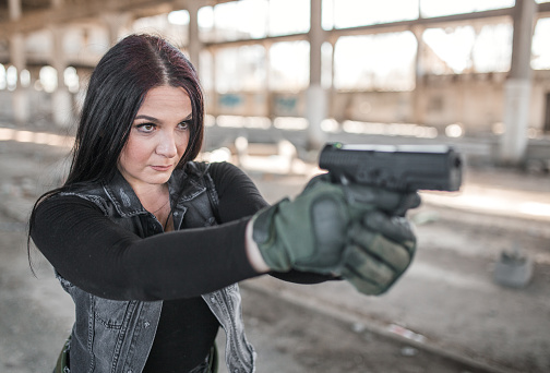Young woman mercenary assassin with gun in war zone in abandoned warehouse