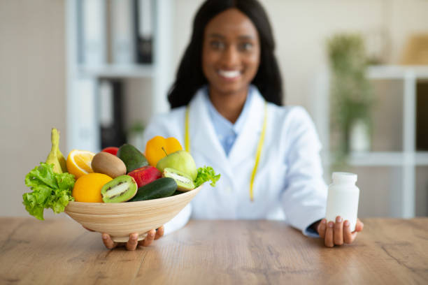 Happy black dietitian offering choice between fresh products and tablets at clinic Happy black dietitian offering choice between fresh products and tablets at clinic. Young nutritionist holding bowl of fruits with veggies and jar of pills or vitamin supplements. Healthy eating fat nutrient photos stock pictures, royalty-free photos & images