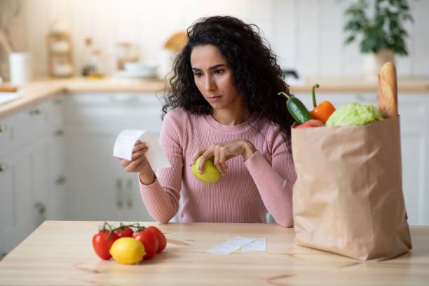 обеспокоенная молодая женщина проверка законопроектов на кухне после продуктовых магазинов - vegies vegetable basket residential structure стоковые фото и изображения