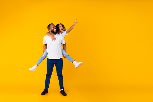 Great Offer. Full Body Length Of Cheerful African American Man Giving Piggy Back Ride For His Daughter, Little Girl Looking Aside And Pointing Fingers Up At Free Copy Space On Yellow Background