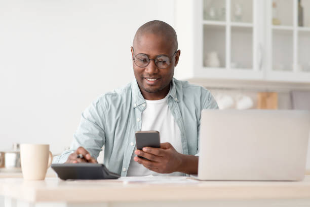 Savings and finances concept. Mature black man using calculator, phone and laptop computer Savings and finances concept. Mature black man using calculator, phone and laptop computer, calculating taxes, sitting in kitchen at home, copy space credit score stock pictures, royalty-free photos & images