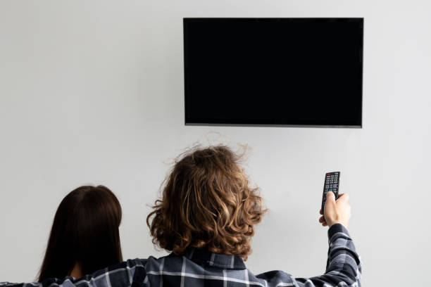 istock Couple Pointing Remote Control On Empty TV Screen Indoor, Back-View 1312618690