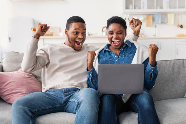 coppia nera che usa laptop che celebra il successo stringendo pugni - shaking fist foto e immagini stock